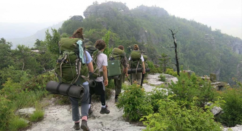 Four students wearing backpacks hike along a rocky trail amid greenery. Based on the rocks and slope ahead of them, the group is in a mountainous landscape.